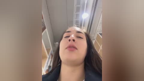 Media: A close-up video of a young woman with fair skin and long brown hair, wearing a dark top, taken from a low angle, showing her from below her chin to her forehead, in an indoor setting with metal beams and white walls.