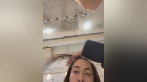 Media: A video captures a woman with light skin and straight brown hair, looking down at a smartphone in a modern, industrial-style room with white walls, exposed pipes, and large arched windows.