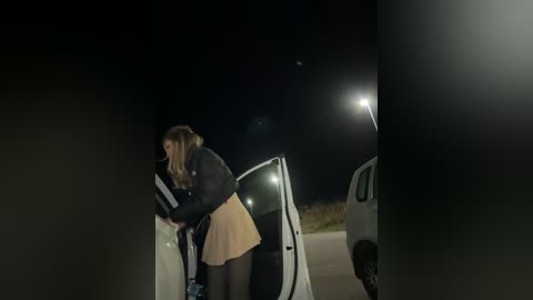 Media: Video of a young woman with long blonde hair, wearing a black jacket and a yellow skirt, loading a white van at night.