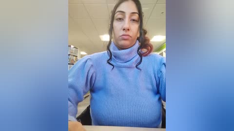 Media: Video of a woman with dark hair, wearing a blue ribbed turtleneck sweater, seated at a desk in a library, with shelves and fluorescent lighting in the background.