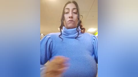 Media: Video of a woman with long dark hair, wearing a light blue turtleneck sweater, captured from a low angle, showing her slightly squinting face. Background is a dimly lit indoor space with white ceiling tiles.