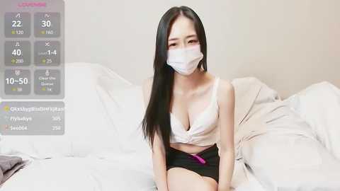 Media: Video of an East Asian woman with long black hair, wearing a white face mask, a pink bra, and black shorts, sitting on a white bed, in a minimalistic bedroom.