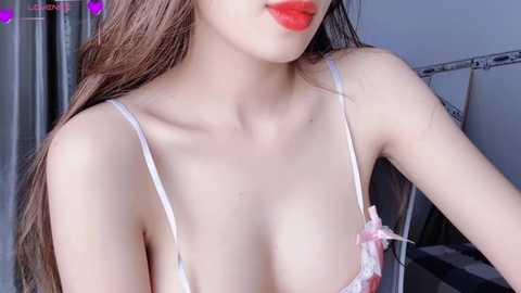 Media: A video of an Asian woman with long brown hair, fair skin, and red lipstick, wearing a white lace bra with pink bows, in a dimly lit bedroom.