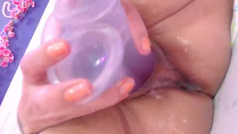 Media: Video of a person's hand with orange-painted nails holding a clear, purple, and green dildo against their wet, glistening, light-skinned genitals in a bathtub.