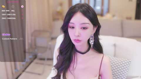 Media: Video of an East Asian woman with long black hair, fair skin, wearing a black choker, and a white dress, sitting on a modern white sofa. Background shows a living room with beige walls and soft lighting.