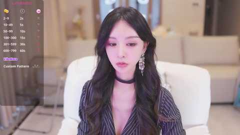 Media: Video of an Asian woman with long black hair, wearing a black pinstripe dress and choker, sitting on a white couch in a modern, neutral-toned living room.