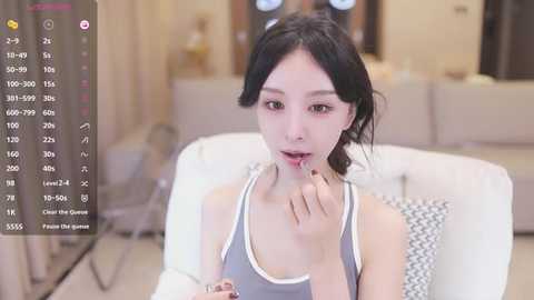 Media: Video of an East Asian woman with pale skin, black hair, and brown eyes, wearing a gray tank top. She licks her finger suggestively, in a modern living room with beige furniture and a TV screen showing a phone app interface.