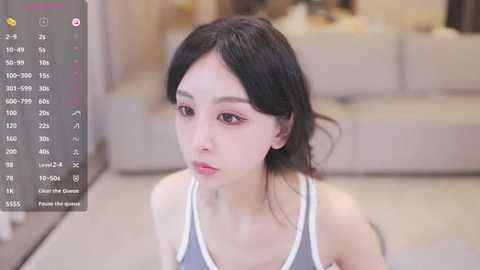 Media: Video of a young East Asian woman with fair skin, black hair, and a neutral expression, wearing a grey tank top, sitting in a beige room with a sofa in the background.