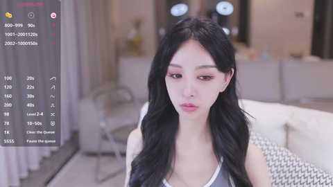 Media: Video of an East Asian woman with long black hair, wearing a gray tank top, in a modern living room with a blurred background.