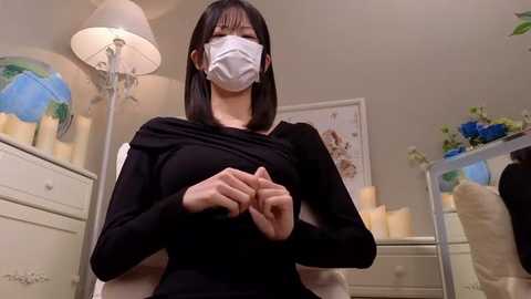 Media: Video of a young woman with a white face mask, black dress, and straight black hair, standing in a softly lit, cozy bedroom with a white dresser, world map, and blue flowers in the background.