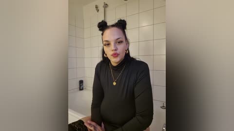 Media: A video of a pregnant woman with light skin, dark hair in two buns, wearing a black long-sleeved top, standing in a tiled bathroom, with a bathtub and showerhead in the background.