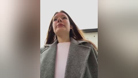 Media: Video of a Caucasian woman with straight, light brown hair, wearing a light gray coat over a white shirt, standing outdoors against a cloudy sky and modern building background.