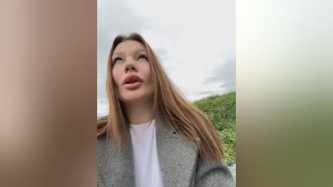 Media: Video of a young woman with long blonde hair, wearing a grey coat and white shirt, looking up at a cloudy sky, with green grass in the background.