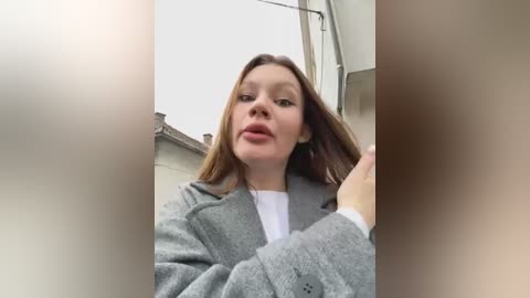 Media: Video of a young woman with fair skin, straight brown hair, wearing a grey cardigan and white shirt, standing outdoors near a building with a sloped roof.