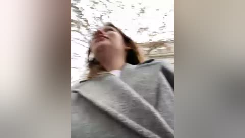 Media: A low-angle video of a woman with light skin and shoulder-length brown hair wearing a gray coat, taken from a blurred, close-up perspective. The background features an overcast sky and blurred tree branches.