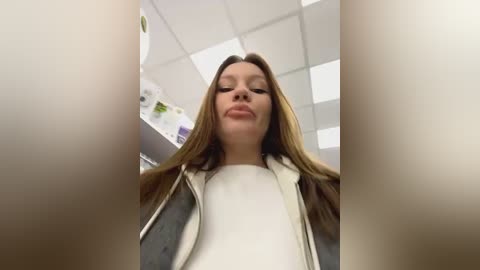 Media: Video of a young woman with straight, light brown hair, wearing a white top and a gray jacket, standing in a white-tiled office with fluorescent lights, looking down at the camera.