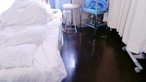 Media: Video of a hospital room with a person in white hospital gown on a bed, dark wooden floor, white curtain, metal stool, and medical equipment.