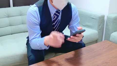 Media: Video of a person in a light blue shirt, dark vest, and striped tie, sitting on a light green couch, texting on a smartphone, wooden table in the foreground.