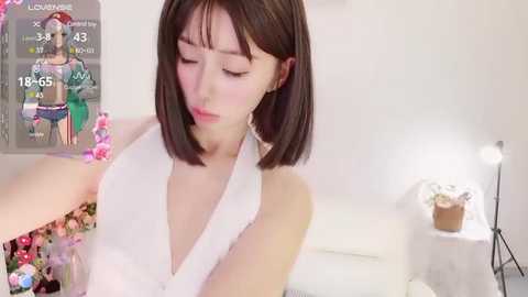 Media: Video of a young Asian woman with straight, shoulder-length brown hair, wearing a white halter top, standing indoors with a virtual reality headset on, next to a potted plant.