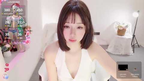 Media: A video of an Asian woman with a short bob haircut, wearing a white halter top, seated in a brightly lit room with a floral arrangement and a digital clock.