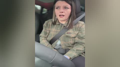 Media: Video of a young woman with straight, shoulder-length brown hair, wearing a green plaid shirt, gray seatbelt, and gray pants, sitting in a car, looking worried.