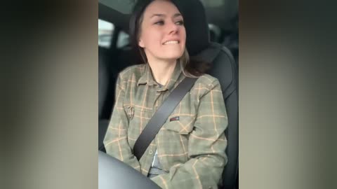 Media: Video of a smiling Caucasian woman with light skin and long brown hair, wearing a plaid shirt, and a seat belt, sitting in a car.