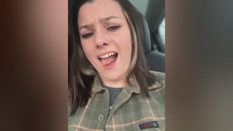 Media: Video of a Caucasian woman with straight, dark brown hair, wearing a green plaid shirt, laughing with her mouth open, sitting in a car.