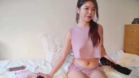 Media: Video of a young Asian woman with long black hair, sitting on a bed in a simple bedroom. She wears a light pink crop top and shorts, holding a phone and a camera.