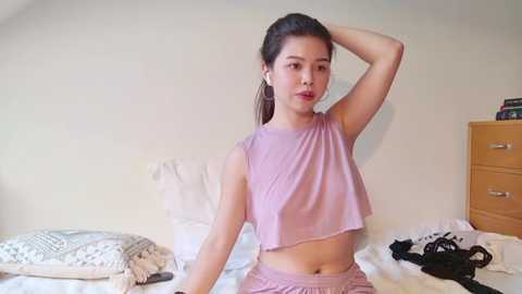 Media: Video of a young Asian woman with light skin, straight brown hair, wearing a pink sleeveless crop top and matching shorts, posing in a messy bedroom with white sheets, a wooden dresser, and scattered clothes.