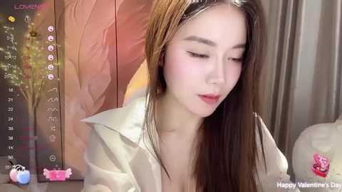 Media: Video of a young Asian woman with long, straight brown hair, fair skin, and closed eyes. She wears a white blouse. Background includes a digital calendar, a pink heart, and a leafy plant.