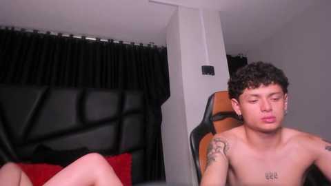 Media: Video of a shirtless, curly-haired, light-skinned man with tattoos, sitting on a red chair with a black headboard, in a bedroom with black curtains.