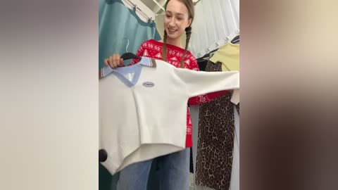 Media: A video of a smiling young woman with light skin, brown hair in braids, wearing a red sweater with white snowflake pattern, holding a white sweater with a blue V-neck, in a clothing store.