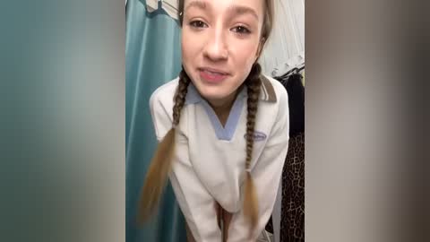 Media: Video of a young woman with light skin and brown braids, wearing a white polo shirt, leaning forward in a bathroom with a teal shower curtain and leopard-print towel in the background.