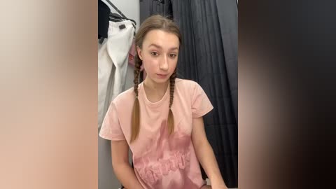 Media: Video of a young, fair-skinned woman with long, light brown pigtails, wearing a pink T-shirt, standing in a dimly lit room with a black curtain and hanging clothes in the background.