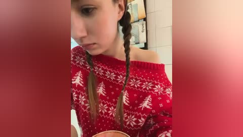 Media: Video of a young woman with light skin and long brown hair in braids, wearing a red sweater with white Christmas tree pattern, sitting in a bathroom with white tiles and toiletries.