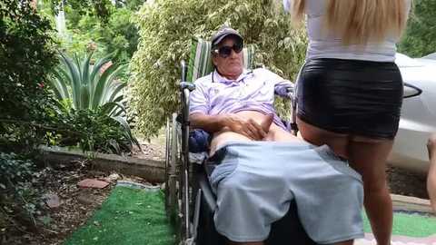 Media: A video captures an older man in a wheelchair with a young woman standing behind him, adjusting his pants. The background features lush greenery and a white car.