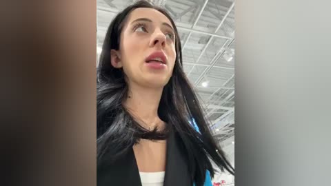 Media: Video of a fair-skinned woman with long black hair and a black blazer, standing in a modern, industrial setting with high ceilings and exposed beams.