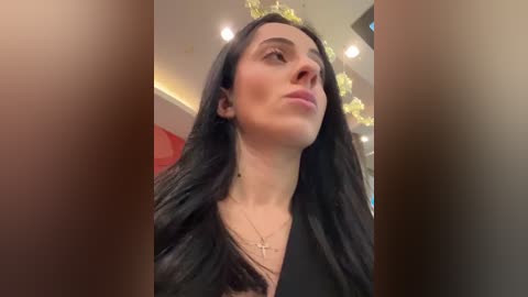 Media: Video of a fair-skinned woman with long, straight black hair, wearing a black top and gold necklace, looking upward with a thoughtful expression. Background features a modern, indoor setting with warm lighting and yellow string lights.