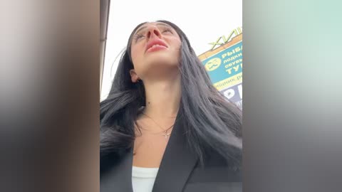 Media: Video of a young woman with long black hair, fair skin, and red lipstick, wearing a black blazer and white top, captured from a low angle, with blurred signs in the background.