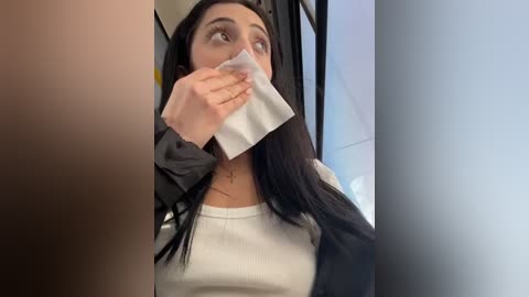 Media: Video of a young woman with long black hair, wearing a white top and black jacket, holding a tissue to her nose, looking upset, in a blurry background.