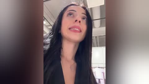 Media: A video of a smiling woman with long black hair, wearing a black top, taken from a low angle, showing her cleavage, in a modern room with white walls and a ceiling fan.