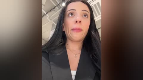 Media: A video of a woman with long black hair, fair skin, and a slightly pained expression, wearing a black blazer and white top, standing indoors against a modern, industrial background.