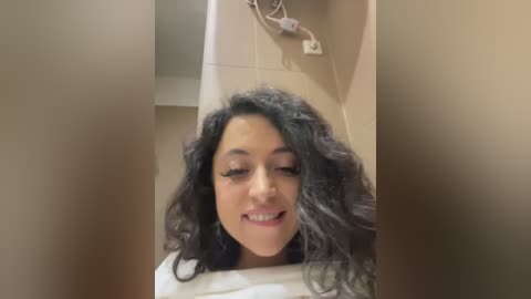 Media: A video of a young woman with curly, dark hair, wearing white clothing, smiling. The background features beige tiles and a light switch.