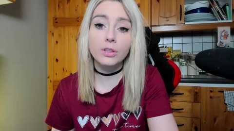 Media: Video of a young blonde woman with fair skin, wearing a red t-shirt with a heart graphic and a black choker, in a wooden kitchen with a countertop and utensils.