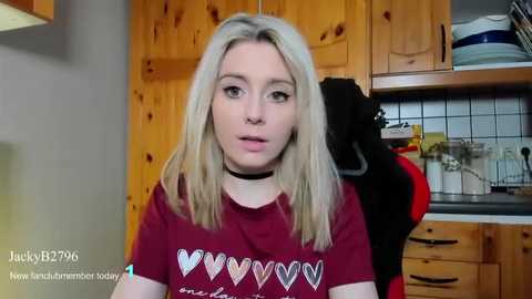 Media: A video of a blonde woman with a choker, wearing a red T-shirt with heart designs, in a wooden kitchen with a black and red jacket hanging on a hook.