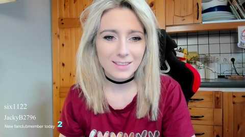 Media: A video of a young blonde woman with a black choker, smiling in a kitchen with wooden cabinets, white tiles, and a red shirt.