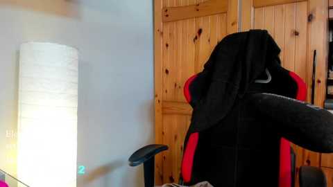 Media: Video of a gaming chair with a black and red backrest, draped with a black cloth, positioned in front of a wooden wardrobe. A cylindrical, white, fabric lamp stands beside the chair. The scene is indoors, likely a bedroom or study.