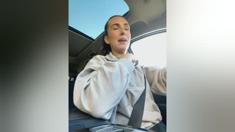 Media: A video of a woman with light skin and dark hair, wearing a grey hoodie, driving a car. She appears relaxed with eyes closed and mouth slightly open. The car interior is visible, with a blue sky visible through the windshield.