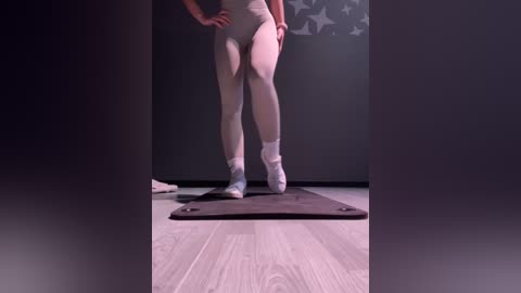 Media: Video of a fit woman in a grey leotard and white socks standing on a yoga mat, with a grey wall and star pattern behind her.