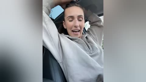 Media: Video of a Caucasian woman with light skin and brown hair, wearing a light gray hoodie, smiling broadly while driving. She has her right arm raised, resting on the car's headrest.